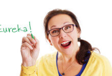 Smiling woman with glasses writes 'Eureka!' in green marker on white background.