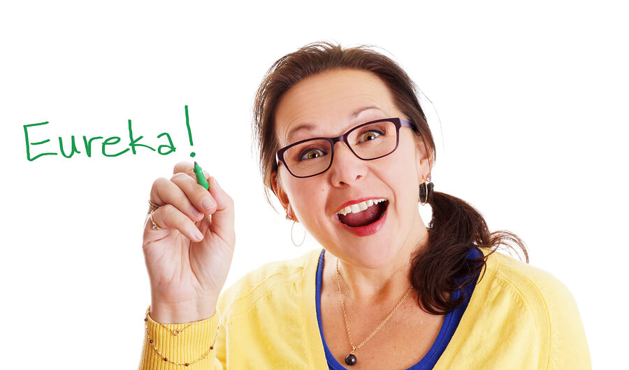 Smiling woman with glasses writes 'Eureka!' in green marker on white background.